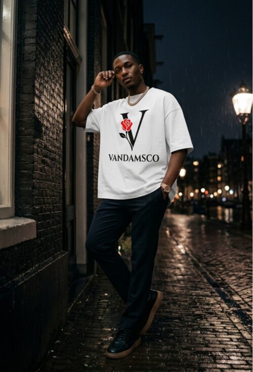 Iemand leunt tegen een bakstenen gevel in een Amsterdamse steeg bij avond in de regen, in een wit oversized Vandamsco T-shirt met de V-roosprint.